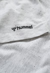 Camiseta blanca con motas negras, presenta un logo negro de "hummel". La tela parece suave con una textura lisa y un diseño casual.