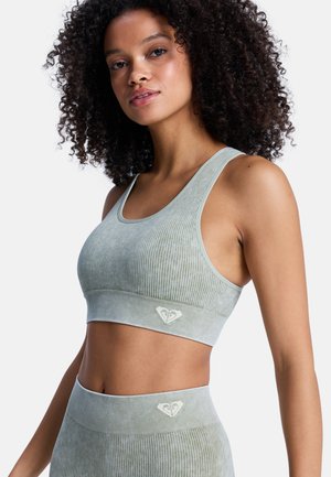 Jonge vrouw met krullend haar, gekleed in een lichtgroene geribbelde sportbh en bijpassende legging met hoge taille, staand tegen een witte achtergrond.