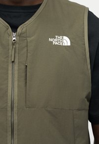 Posnetek v bližini osebe, ki nosi olivno zelen brezrokavnik z zadrgo spredaj in belim logotipom "The North Face" na prsih.