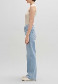 Mujer vistiendo un top de punto beige, jeans rectos de tiro alto de color azul claro y zapatillas blancas, de pie de perfil sobre un fondo liso.