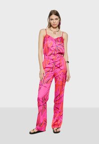 Roze satijnen outfit met een bloemenpatroon. Bestaat uit een mouwloos topje en een broek met wijde pijpen, aangevuld met zwarte teenslippers en eenvoudige sieraden.