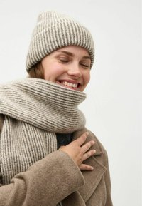 Beige gebreide muts en bijpassende sjaal met verticale strepen, gemaakt van zachte, gestructureerde wol. Gelaagd met een bruine jas en een zwarte top eronder.