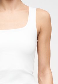 Γυναίκα που φοράει λευκό αμάνικο τοπ, με εμφανές το πάνω μέρος του κορμού και το αριστερό χέρι της, πάνω σε απλό φωτεινό φόντο.