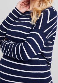 Femme blonde portant un pull à manches longues rayé bleu marine et blanc, avec un bras croisé et la main touchant son visage.