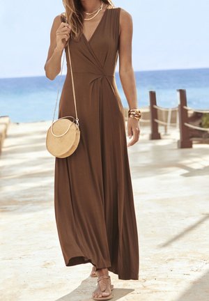 Femme marchant sur un sentier de plage ensoleillé, vêtue d'une robe maxi marron sans manches, de sandales beige, d'un sac rond en paille et de bijoux en or.