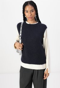 Suéter sem mangas de malha azul-marinho sobre uma blusa de manga longa creme, combinado com calças largas cinza-escuro e uma alça de bolsa prateada.