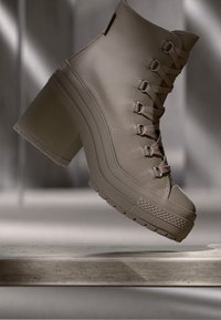 Beige ankle boot with a high block heel, featuring a textured leather finish, lace-up design, and prominent rubber sole with patterns.