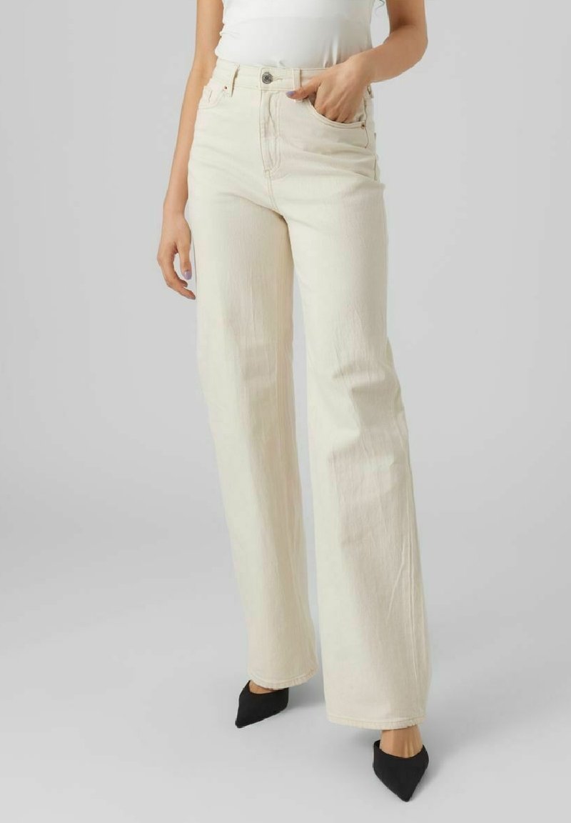 Femme portant un jean large crème taille haute, un top blanc et des escarpins noirs à bout pointu, main dans la poche.