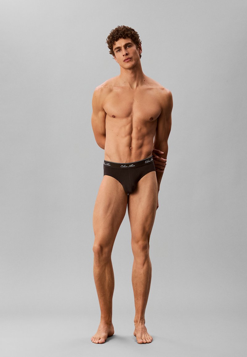 Jeune homme musclé debout pieds nus portant un sous-vêtement noir avec une ceinture de marque, posant les mains dans le dos sur un fond gris uni.