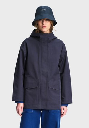 Femme portant une veste à capuche bleu marine foncé, un chapeau bob assorti avec le logo "AIGLE" et un pantalon bleu, sur un fond clair uni.