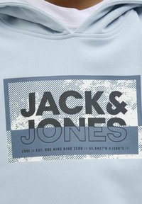 Felpa blu chiaro con cappuccio, caratterizzata da una grafica rettangolare grigia che mostra il testo "JACK & JONES" con un motivo a strisce e coordinate geografiche.