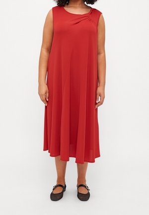 Mujer con un vestido midi rojo sin mangas y de corte holgado, con detalles fruncidos en el escote, combinado con zapatos negros de hebilla.