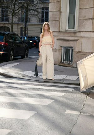 Mujer joven con camiseta beige clara y pantalones anchos de pie en un paso de peatones urbano, sosteniendo una bolsa grande con borlas cerca de un edificio de piedra y coches estacionados.