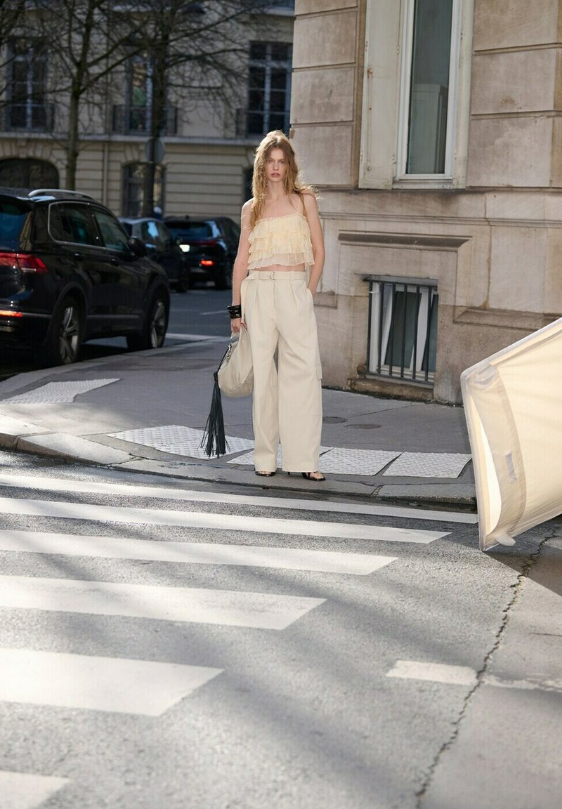 Mladá žena v světle béžovém topu a širokých kalhotách stojí na městském přechodu pro chodce, drží velkou tašku s třásněmi u kamenné budovy a zaparkovaných aut.