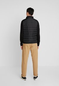 Czarna pikowana kamizelka z wysokim kołnierzem i kieszeniami po bokach, noszona na czarnej koszuli z długim rękawem i beżowych spodniach. Czarne tenisówki.