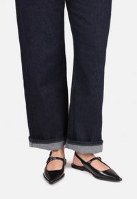 Zapatos slingback negros, con punta afilada y suela plana, que presentan un detalle de hebilla y una parte trasera abierta, combinados con jeans de mezclilla oscura remangados.