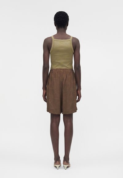 Άτομο που στέκεται με την πλάτη, φοράει ελιάς πράσινο αμάνικο μπλουζάκι, καφέ σορτς με σχέδια και μπεζ πέδιλα με τακούνι σε ένα μονόχρωμο φόντο.