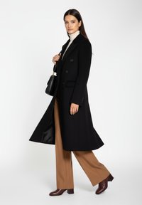 Sort lang frakke med skræddersyet pasform, der har dobbeltradede knapper og sidelommer. Bæres med beige bukser og bordeaux farvede hæle.