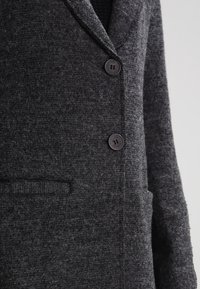 Cappotto grigio testurizzato con colletto a bavero, due bottoni frontali e una tasca verticale con un motivo sottile. Materiale morbido simile alla lana.