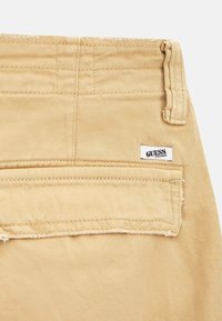 Pantalon en denim beige avec une poche arrière portant une étiquette blanche indiquant "GUESS Originals". Tissu texturé et détails de couture visibles.
