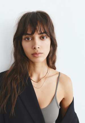 Jeune femme aux longs cheveux foncés, vêtue d'un débardeur gris sous un blazer noir, accessoirisée de deux fines chaînes en or, sur un fond blanc uni.