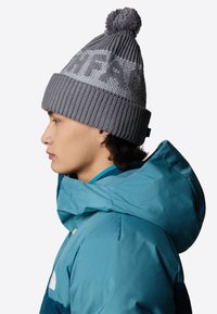 Beanie grigia lavorata a maglia con un grande pom-pom sulla parte superiore, caratterizzata da un motivo testurizzato e un polsino a costine, indossata da una persona in giacca teal.