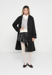Fiatal nő áll, egyik kezével a csípőjén, fekete trench coat-ban, mintás pulóver mellényben, fekete bőr szoknyában, fehér harisnyában és sötét balettcipőben.