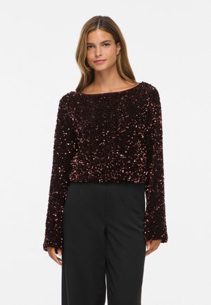 Modèle portant un haut à manches longues à sequins, de couleur bordeaux foncé, et un pantalon noir, devant un fond blanc uni.
