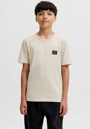 Junge mit kurzen dunklen Haaren, der ein beiges kurzärmliges Hemd mit einem kleinen schwarzen Fleck und schwarze Hosen trägt, steht vor einem einfarbigen Hintergrund.