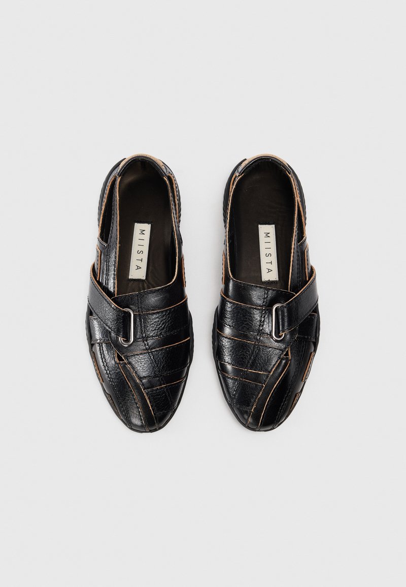 Scarpe di pelle nera con un design incrociato, caratterizzate da cuciture marroni e una fascia decorativa con fibbia in metallo che attraversa il collo del piede.