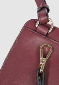 Angolo di borsa a mano in pelle color bordeaux con ferrature dorate, caratterizzata da una finitura testurizzata, dettagli neri e una chiusura con moschettone rimovibile.