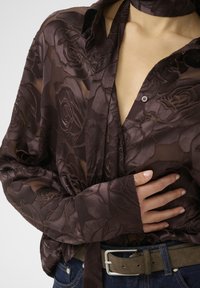 Blusa trasparente marrone con motivo a rose, chiusura frontale con bottoni, maniche lunghe e dettaglio del collo legato, abbinata a jeans casual e una cintura grigia.
