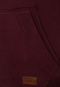 Sudadera burdeos con bolsillo frontal, material de algodón suave, costuras visibles y una pequeña etiqueta de cuero en la parte inferior izquierda.