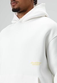 Sweat à capuche blanc en tissu doux, doté d'une poche kangourou et d'un logo jaune subtil brodé sur la poitrine.