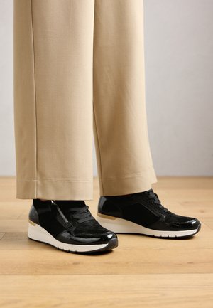 Černé wedgové tenisky s detaily na zipu, které jsou nošeny s béžovými širokými kalhotami na dřevěné podlaze.