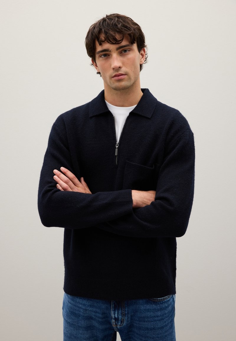 Pullover a maglia navy con colletto con zip, dotato di tasca frontale, texture morbida e vestibilità rilassata, abbinato a jeans azzurri chiari.