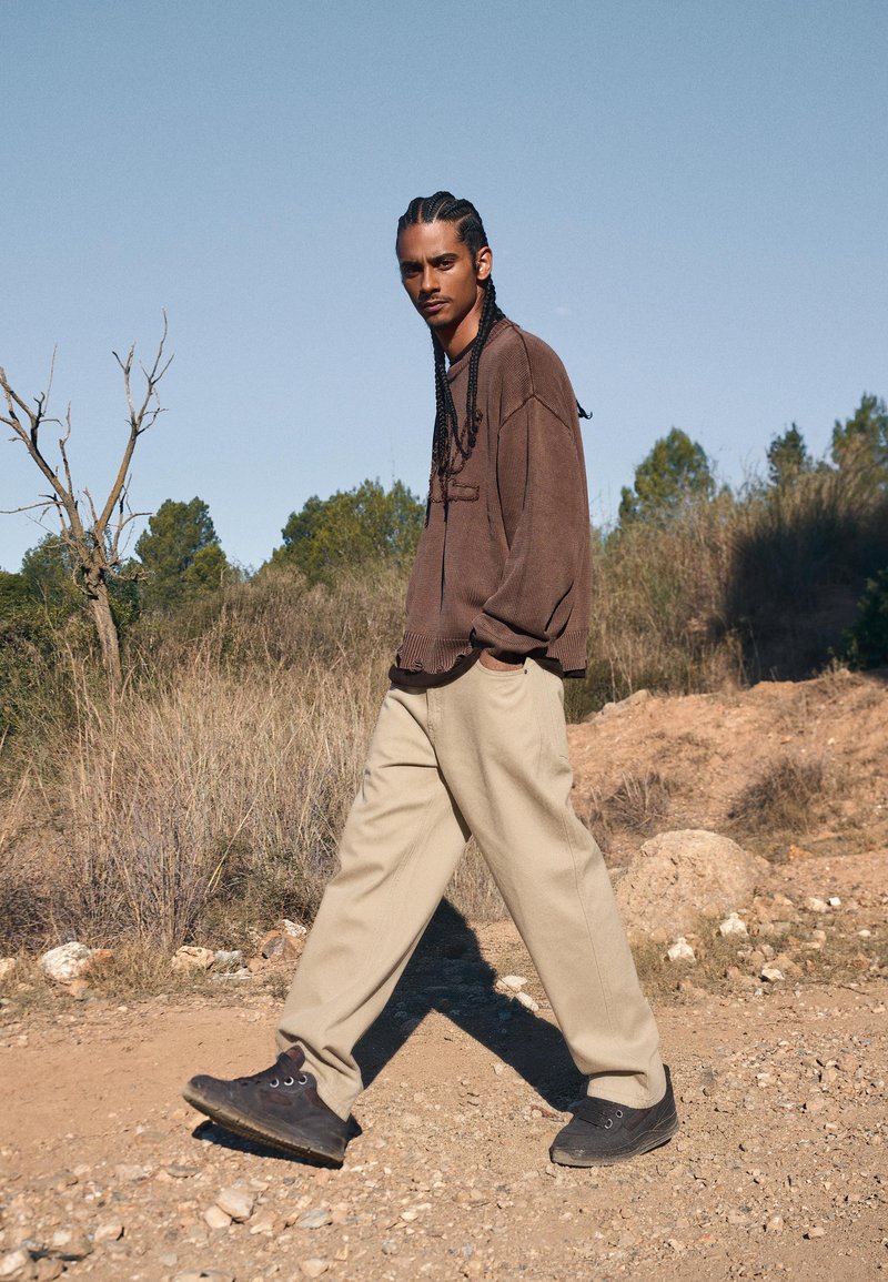 Brun stickad tröja, beige tapered byxor och svarta hög-upp sneakers. Flätad frisyr, står på en grusväg med en naturskön landskap.