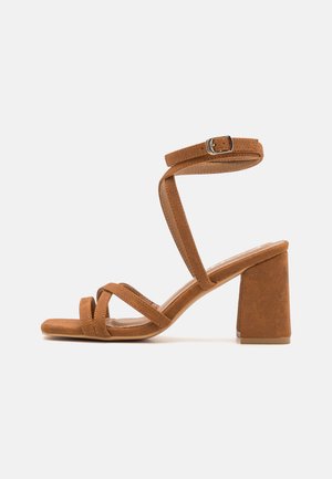 Sandales à talons en daim synthétique marron avec un large talon bloc, plusieurs brides croisées et une boucle ajustable à la cheville. Texture douce, design à bout ouvert.