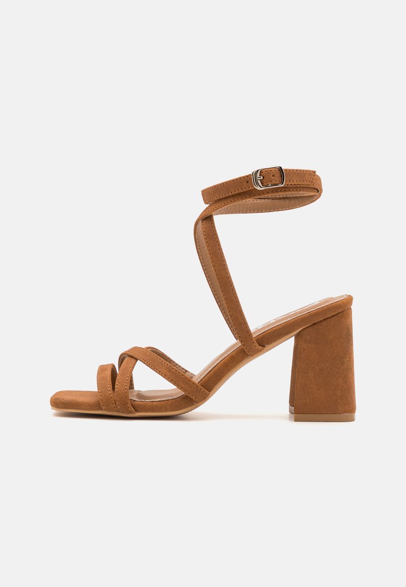 Sandales à talons en daim synthétique marron avec un large talon bloc, plusieurs brides croisées et une boucle ajustable à la cheville. Texture douce, design à bout ouvert.