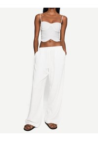 Bílý zkrácený top s volánkovým lemem a tenkými ramínky, kombinovaný s volnými bílými kalhotami s elastickým pasem a kapsami. Hnědé sandály.