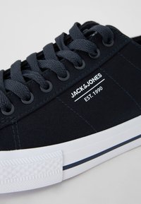 Námořnické plátěné tenisky s bílou gumovou podrážkou, kulatou špičkou a šněrovacím designem. Na boku je vytištěno 'JACK & JONES' a rok založení.