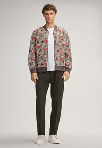 Giacca bomber con motivo di labbra rosse e linee ondulate, abbinata a una t-shirt bianca e pantaloni verde scuro. Sneakers bianche completano il look.