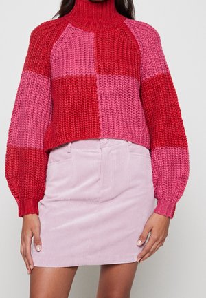 Femme portant un pull à col roulé en maille épaisse rouge et rose, associé à une mini-jupe en velours côtelé rose clair sur un fond uni.