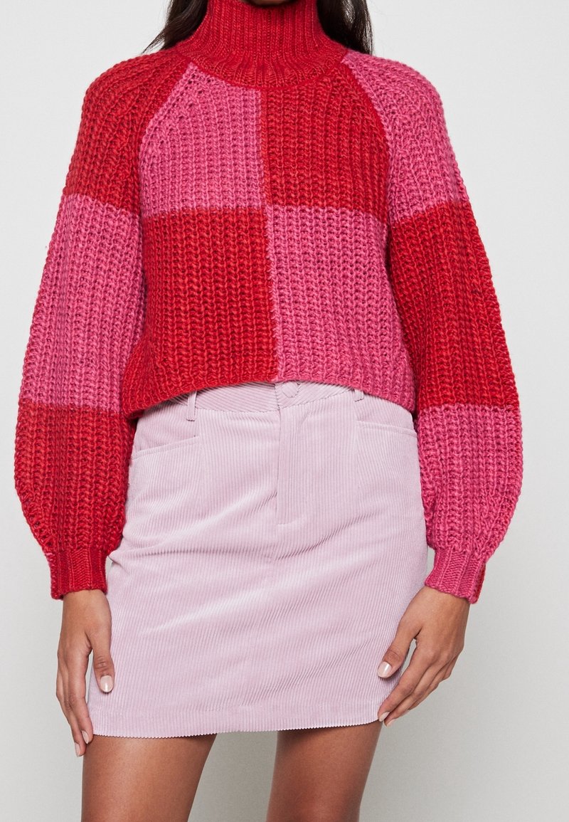 Femme portant un pull à col roulé en maille épaisse rouge et rose, associé à une mini-jupe en velours côtelé rose clair sur un fond uni.