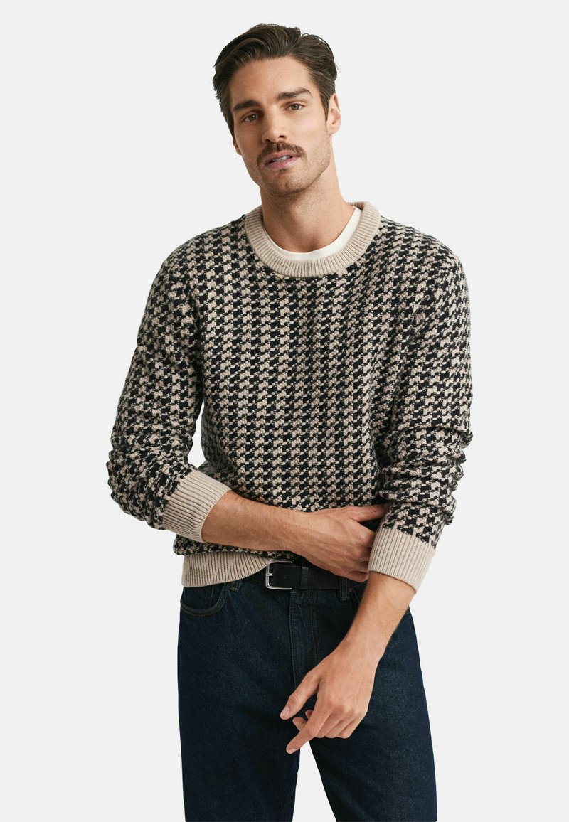 Mann, der einen schwarzen und beigen Hahnentritt-Pullover mit beigen Bündchen und Kragen trägt, kombiniert mit dunkelblauen Jeans und einem schwarzen Gürtel, posiert vor einem weißen Hintergrund.