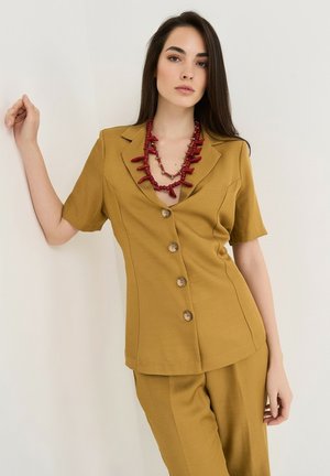 Femme appuyée contre un mur portant un blazer moutarde à manches courtes boutonné, un pantalon assorti et un collier rouge audacieux.