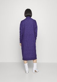 Lång lila tröjklänning gjord av texturerad tyg, med hög halsringning och långa ärmar, matchad med vita klackboots.