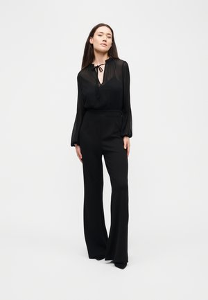 Une femme porte un chemisier noir transparent à nouer au cou, un pantalon à jambes larges et des chaussures noires sur un fond clair et uni.