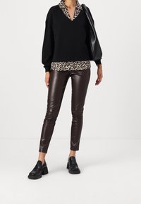 Donna che indossa un maglione nero sopra una blusa con stampa leopardata, pantaloni in pelle marrone scuro, mocassini neri spessi e porta una borsa a tracolla nera.