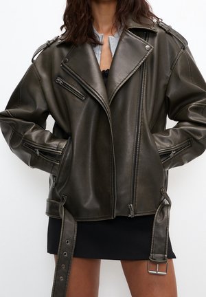 Mujer que lleva una chaqueta de cuero de motorista marrón oscuro de gran tamaño con múltiples cremalleras y correas con cinturón, combinada con una minifalda negra.